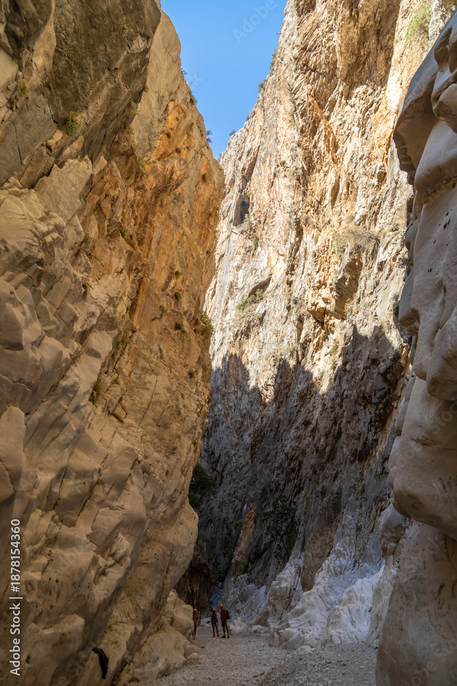 Obraz premium Low angle view of massive limestone cliffs in Saklikent Canyon. Tiny silhouettes of hikers walking between high steep walls of mountain gorge. September, Fethiye, Turkey.