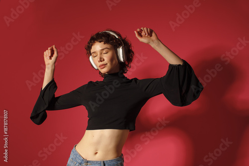Young woman dancing and listening to music with headphones on red background. Freedom, joy, lifestyle, self expression and music concept.