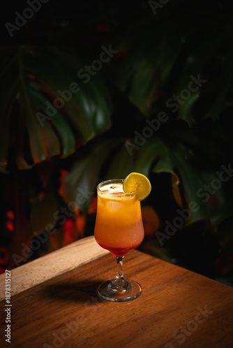 Exotic Tropical Layered Cocktail with Lime Garnish on a Wooden Table against Dark Foliage
