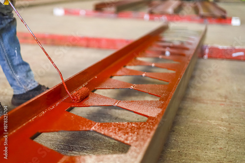 Worker is painting large metal beam with red paint, showcasing industrial craftsmanship. beam features lattice design, and scene captures focus on detail and precision