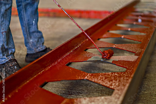 Person is painting metal beam with red paint using brush. beam has series of triangular cutouts and is placed on concrete floor. person is wearing jeans and black shoes, focusing on task
