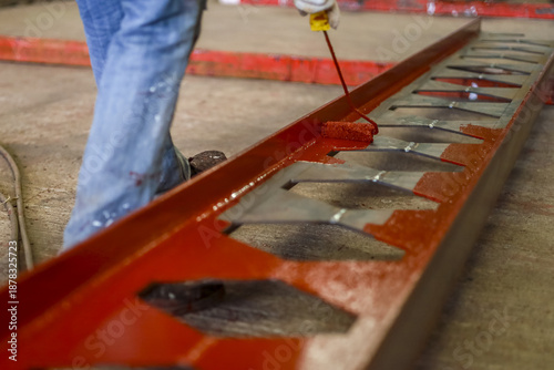 Person is painting metal structure with red paint using roller, focusing on even coverage. scene suggests industrial work, highlighting importance of maintenance and craftsmanship
