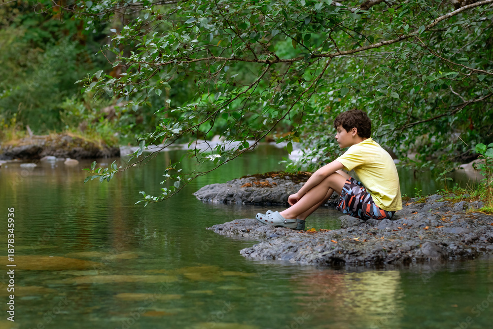 Fototapeta premium Teenager boy is sitting on a big rock in the mountain rocky river with clear green waters in summer forest. Scene under branches with green lush foliag