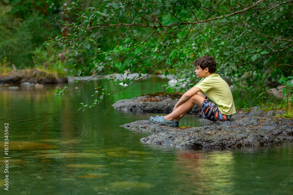 Fototapeta premium Teenager boy is sitting on a big rock in the mountain rocky river with clear green waters in summer forest. Scene under branches with green lush foliag