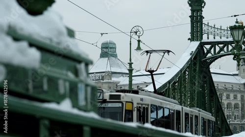 Close-up view of Liberty Bridge in Budapest, Hungary, as a tram moves across during winter. Snow-covered structure, cold weather, and public transportation create a calm urban winter atmosphere.