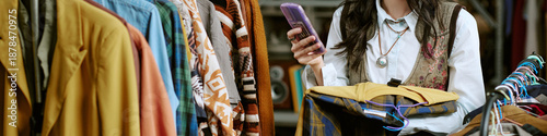 Photos Unrecognizable woman browsing second hand clothing rack, holding smartphone in o