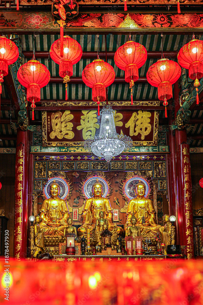 Fototapeta premium Golden Buddha statues inside Wat Mangkon Kamalawat, Bangkok Chinatown. Soft blurred worship candles in foreground and red lanterns above create a peaceful Lunar New Year spiritual atmosphere.