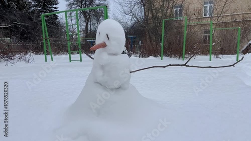 A sad snowman with a carrot nose in a snowdrift with branches in his arms against the backdrop of old houses