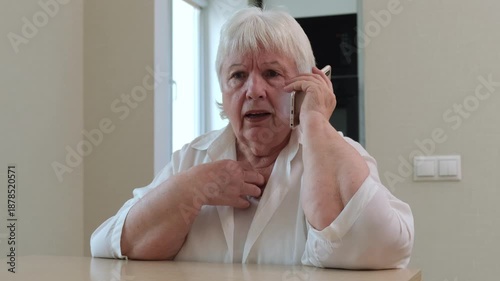 Elderly woman sitting at table and emotional speaking on mobile phone. Concept of news, stress and call from scammer