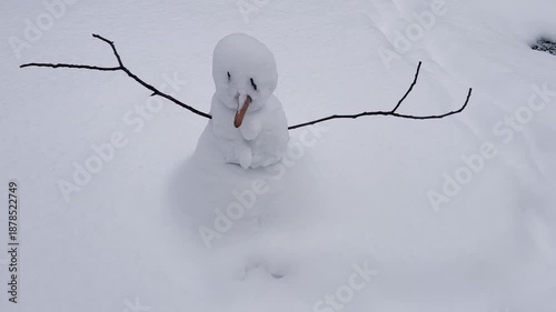 A sad snowman with a carrot nose in a snowdrift with branches in his arms against the backdrop of old houses