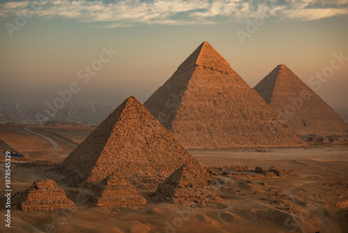 Aerial view of the majestic pyramids of Giza stand resolute against the soft, hazy horizon, their ancient stones glowing with the warmth of the setting sun, Giza, Egypt.