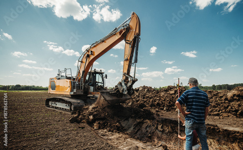 Excavator digging and shaping trench in farmland while worker stands with measuring rod. Heavy machinery earthworks for drainage or pipeline installation. Rural construction scene under blue sky.