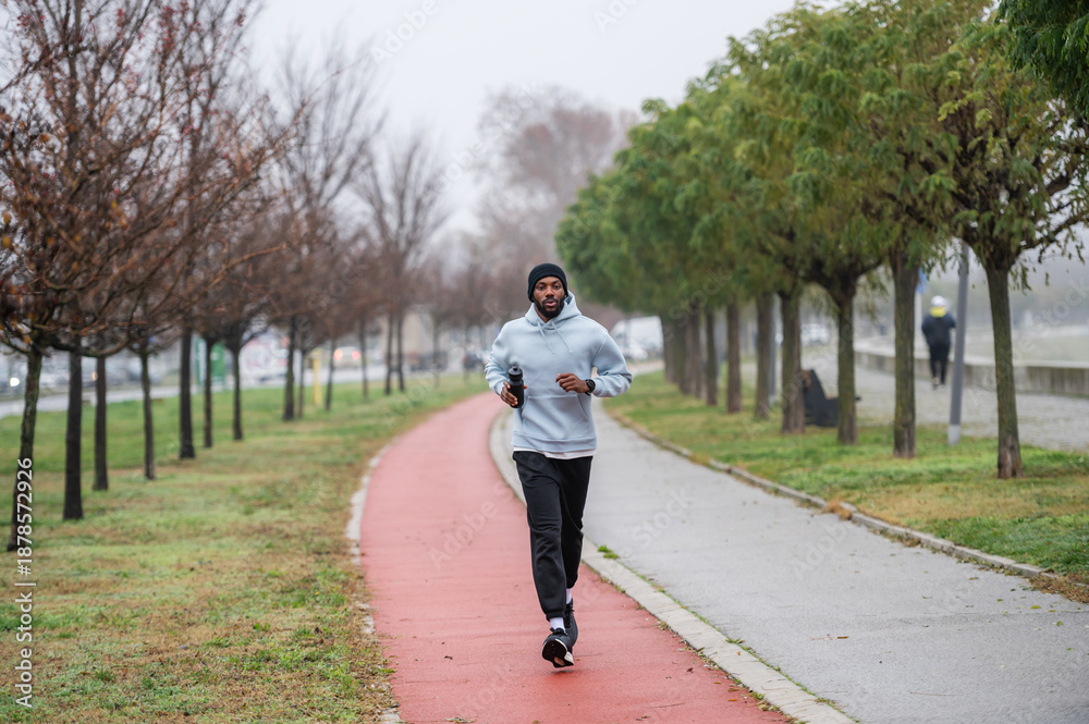 Fototapeta premium Man jogging on a red running track in a misty park, wearing a hoodie and sportswear. Outdoor fitness, healthy lifestyle, and endurance training on a foggy autumn or winter morning