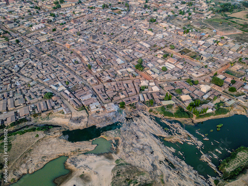 Aerial view of a vibrant urban sprawl meeting the rugged terrain and tranquil waters of Kabala Costain, Kaduna, Nigeria.
