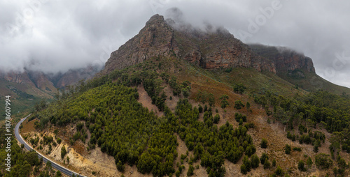 Aerial view of a winding road snakes along the base of a majestic mountain, its peak shrouded in mist, revealing a landscape of contrasting textures and tones, Paarl, Western Cape, South Africa.