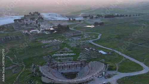 Aerial drone footage of the ancient Roman amphitheater of Hierapolis overlooking Pamukkale, Turkey, at sunset. The well-preserved stone structure stands on a hillside above the famous white travertine