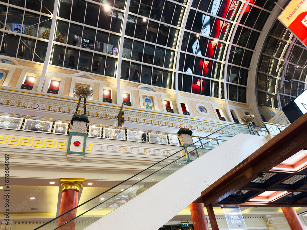 Fototapeta premium Elegant glass atrium mall interior in Manchester with escalator, ornate decor, and bright skylight