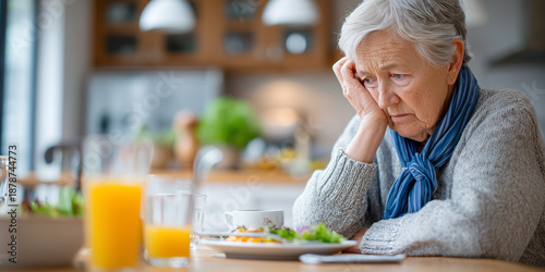 Lonely elderly woman with gray hair looking sad and tired while sitting at a table with untouched food in a cozy kitchen.