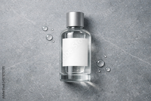 A clear glass bottle with a silver cap and blank label surrounded by water droplets on a grey stone surface viewed from directly above