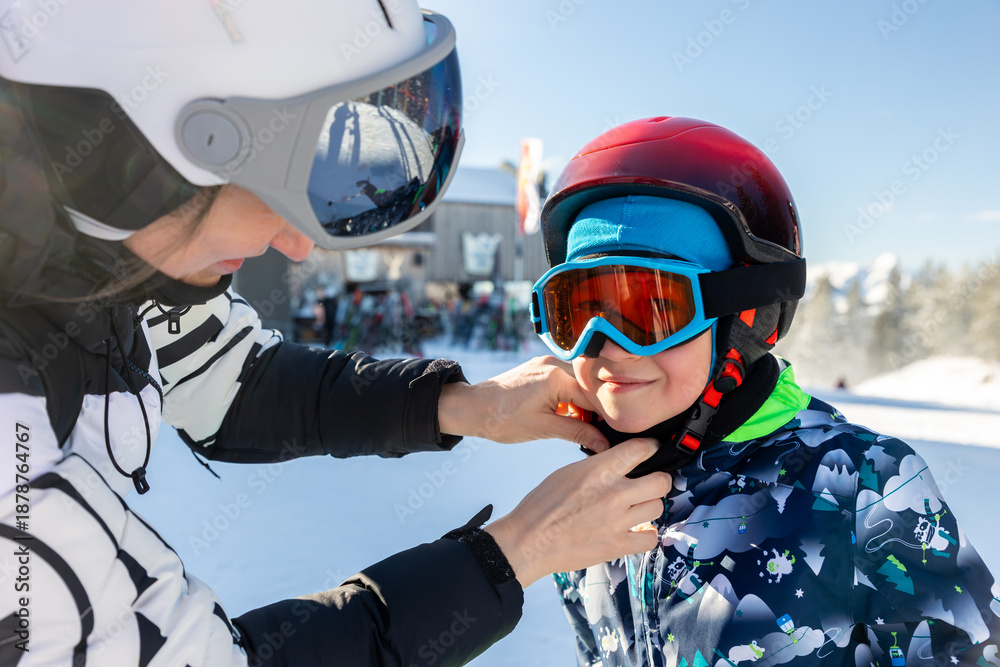 Fototapeta premium Mother helps son fasten ski helmet before slope activity at alpine resort. Winter family moment shows safety, care, bonding snow sport lifestyle protective gear outdoor recreation, childhood learning