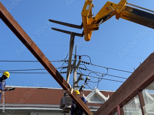 Yellow hydraulic forklift arm stabilizing a long wooden beam close to overhead power lines, highlighting construction risk, safety concerns, and urban infrastructure work.