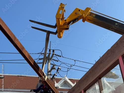 Yellow hydraulic crane fork lifting a long wooden beam near overhead power lines during building renovation, highlighting construction risk, heavy machinery operation, and workplace safety.