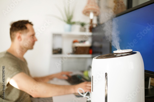 Bilde på lerret Modern air humidifier device during sitting workplace, young man using computer work on internet, enjoying aromatherapy steam scent from essential oil diffuser in room at home office