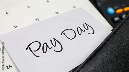 Handwritten words PAY DAY on a white note placed on a calendar with a calculator and leather folder.