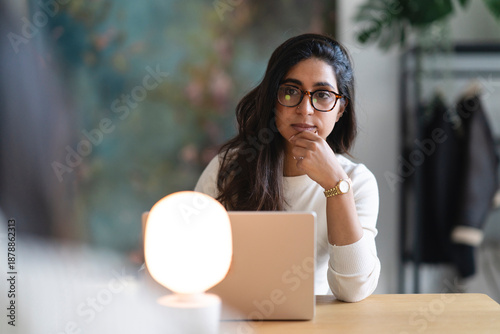 A woman wearing glasses sits at a desk with a laptop and a lamp, deep in thought as she works.