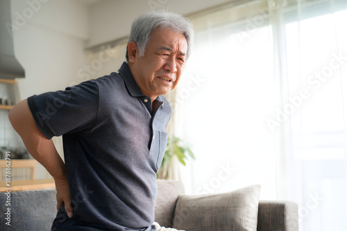 Elderly man struggling to stand up from sofa due to back pain. Senior citizen suffering from chronic joint pain in living room. Daily life struggle.