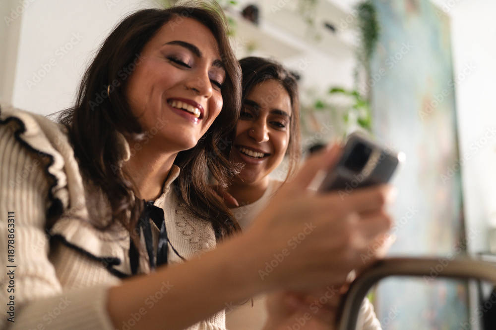 © Studio Marmellata - Two women share a laugh while looking at a smartphone together, enjoying a moment of connection and shared amusement.