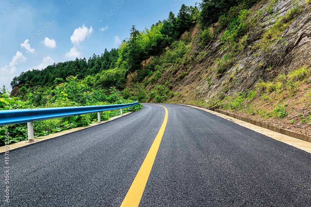 Fototapeta premium Scenic asphalt road winding through green mountains under a bright blue sky.
