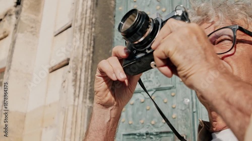 Caucasian senior man with gray curly hair taking photo with camera outdoors. Male photographer capturing architecture in sunlight. Close up side view of aged man shooting urban scenery.
