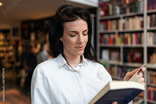 Photos Smiling woman in a white shirt holds a book while browsing shelves in a modern bookstore with rows of colorful books, shot indoors with shallow depth of field for retail and education concepts