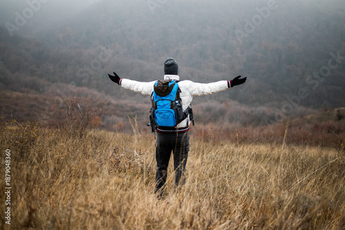 Rear view of hiker in warm clothing standing in nature with backpack and arms outstretched, enjoying freedom and achievement in a foggy winter landscape.