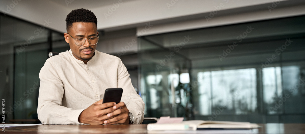 Naklejka premium Busy African American entrepreneur using cell phone working in office. Professional young Black business man holding mobile cellphone looking at cellphone making financial bank payment at work.