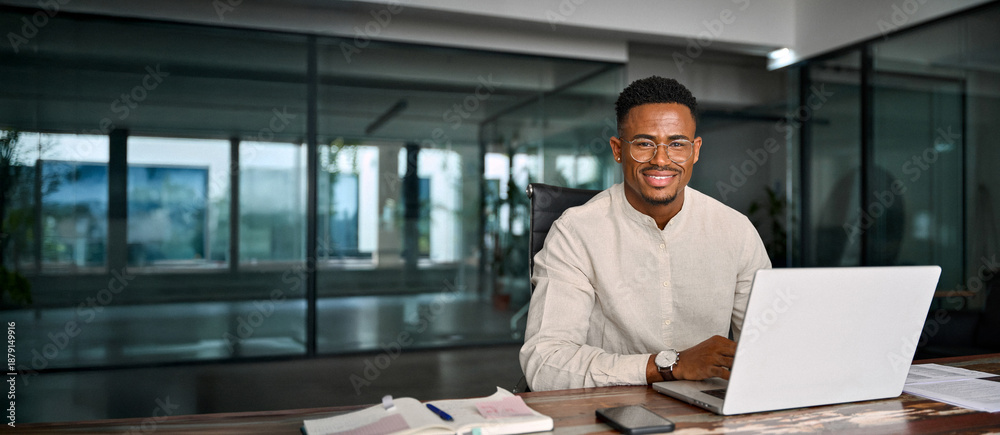 Obraz premium Portrait of smiling male company employee or entrepreneur in office. Happy young professional African American business man in glasses looking at camera sitting at work desk using computer.