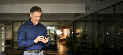 Smiling mature business man corporate leader using cellphone standing in office. Happy busy middle aged older male entrepreneur holding mobile phone tech device looking at smartphone at work.