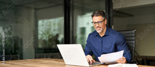 Happy busy older professional businessman entrepreneur using laptop holding bill corporate document in office. 50 years old man executive manager or business owner reading paper working on computer