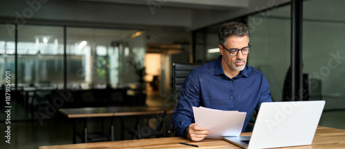 Photos Middle aged professional business man using laptop computer reading document at work