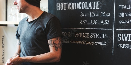 Tablou pe pânză Caucasian man in casual attire with tattoos stands by a chalkboard menu