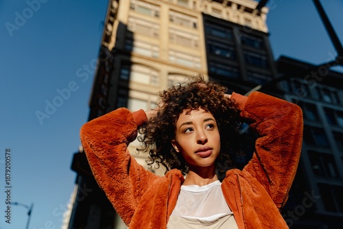 Fotografie Woman in orange jacket stands in sunlight, urban setting