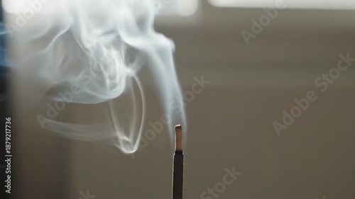Wisps of White Incense Smoke Curl and Rise from a Dark Stick Against a Softly Blurred Neutral Background in Natural Light