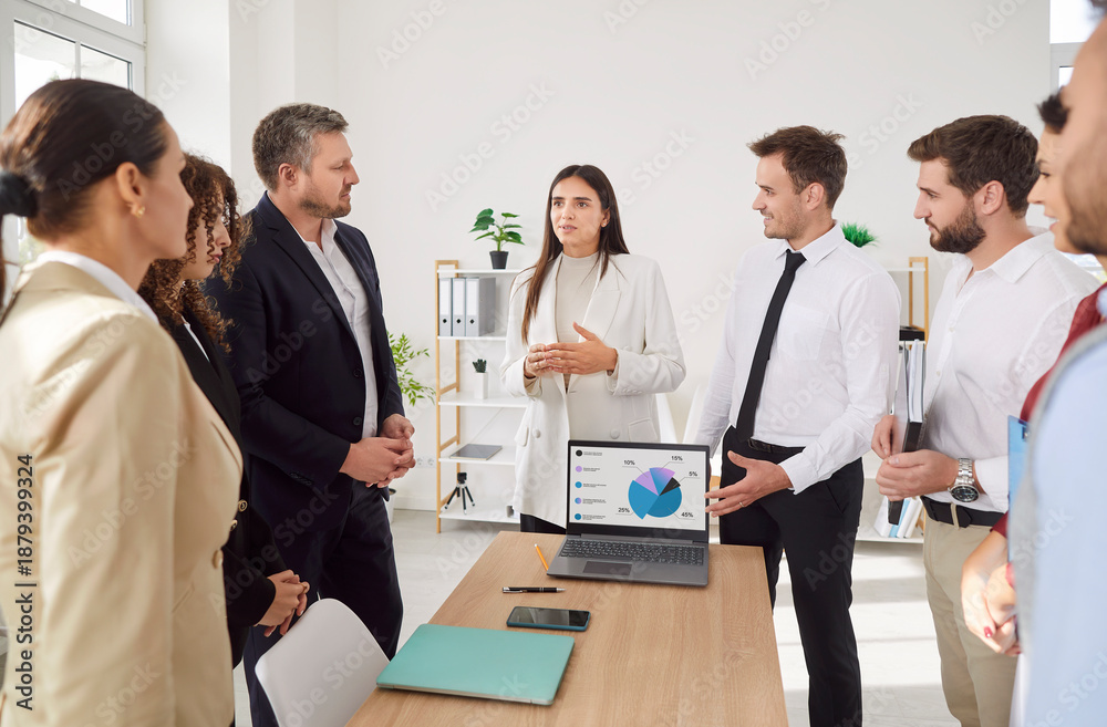Fototapeta premium Young confident business people discussing work project and company growth on laptop with a group of company employees standing in meeting room during job conference in modern office.