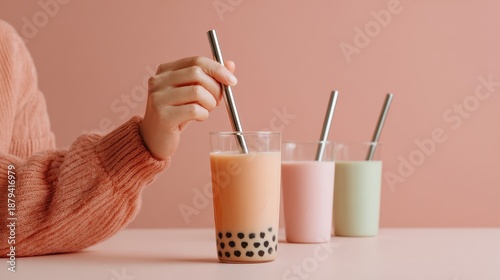 Hand holding a metal straw in a glass of bubble tea with colorful drinks on a pink background, showcasing a trendy beverage experience