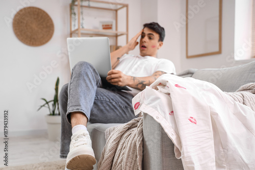 Canvas Print Shocked man with laptop and kiss marks on his shirt on sofa at home