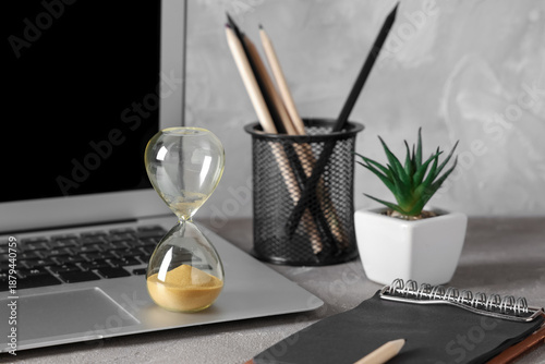 Stylish hourglass with yellow sand, stationery and laptop on table at workplace