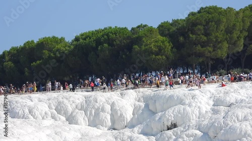 Visitors walk across white calcium carbonate travertines of Pamukkale cotton castle landscape. Tourists explore shimmering mineral terraces formed by hot spring waters at world heritage site.