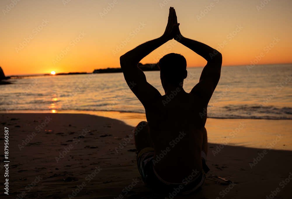 Fototapeta premium Dark silhouette is sitting cross-legged and meditating on sandy beach at sunset, sandals nearby