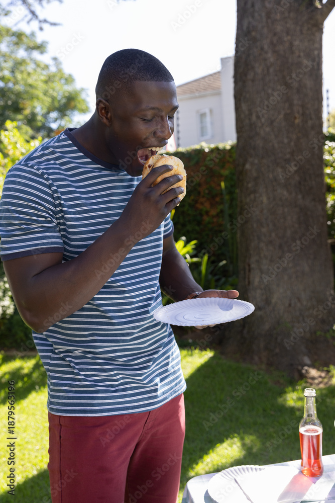 Fototapeta premium African american man standing on lawn eating hamburger from paper plate by table in backyard